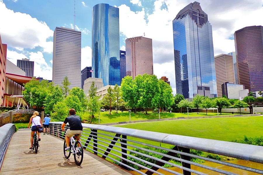 About Our Agency - People Bike Across a Wooden Bridge in a Park, the Houston Skyline Overhead, on a Sunny Day